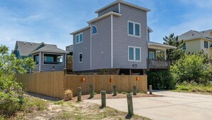 A two-story residential building with a flat roof, a balcony on the second floor, and a wooden fence in the front yard. - Shore Thing - Corolla, Ocean Sands, 60 Yards to Beach, Ocean View, Pool, Hot Tub, Pets OK (Corolla)