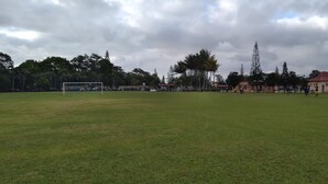 Un extenso campo cubierto de hierba con un arco, rodeado de árboles y un edificio de techo rojo.