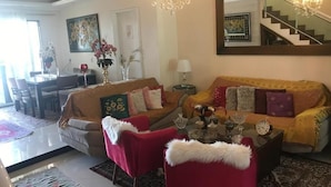Een woonkamer met een bank, fauteuils, een salontafel en een kroonluchter. Op de achtergrond is een zithoek met een tafel en stoelen.