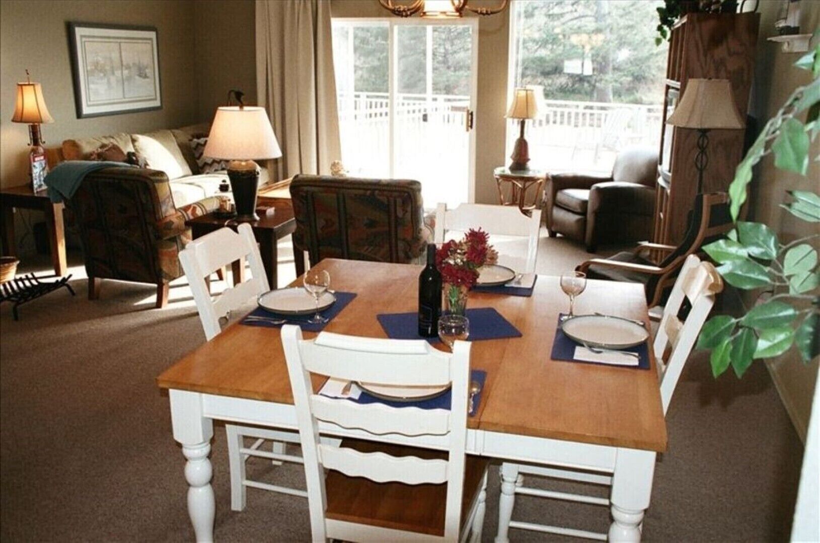 Un coin repas avec une table en bois dressée pour quatre personnes, entourée de chaises blanches. On y trouve deux petites tables d’appoint avec des lampes, ainsi qu’une fenêtre donnant sur la vue d’arbres.