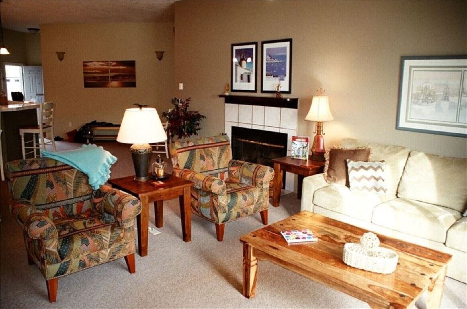 A living room with patterned armchairs, a wooden coffee table, and a sofa. There is a fireplace with framed pictures above it.