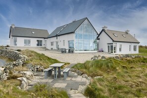 Ein modernes, zweistöckiges Haus mit einer großen Glasfassade, einem steinbelagten Weg, der zu einer Sitzgelegenheit führt, und einer grünen Landschaft.
