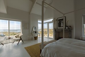 Une chambre avec un grand lit, un coin salon avec un fauteuil et une table, une cheminée et une vue sur la plage.