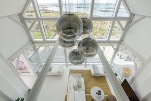 A high-ceiling room with a large window overlooking a coastal landscape, featuring a white sofa, a round table, and hanging spherical light fixtures.