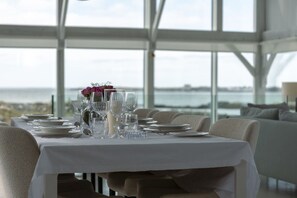 Une table à manger dressée pour six personnes, avec vue sur la mer grâce à de grandes fenêtres.