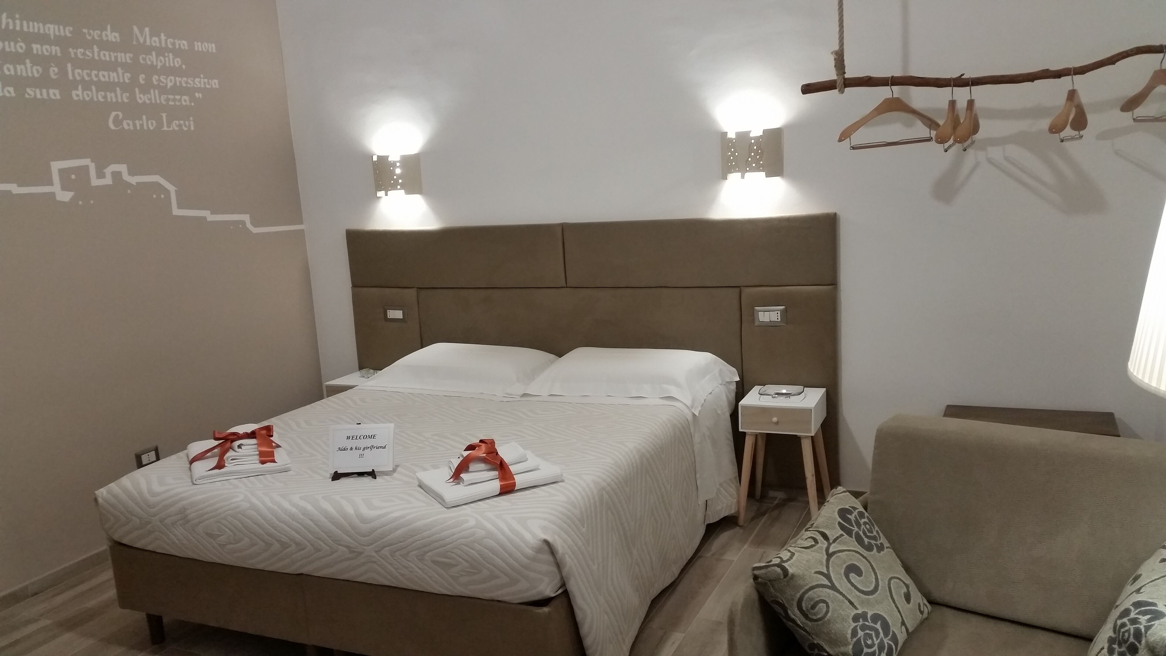 Ein ordentlich bezogenes Bett mit einer Willkommensanzeige, ein Nachttisch mit Lampe und ein Kleiderständer.