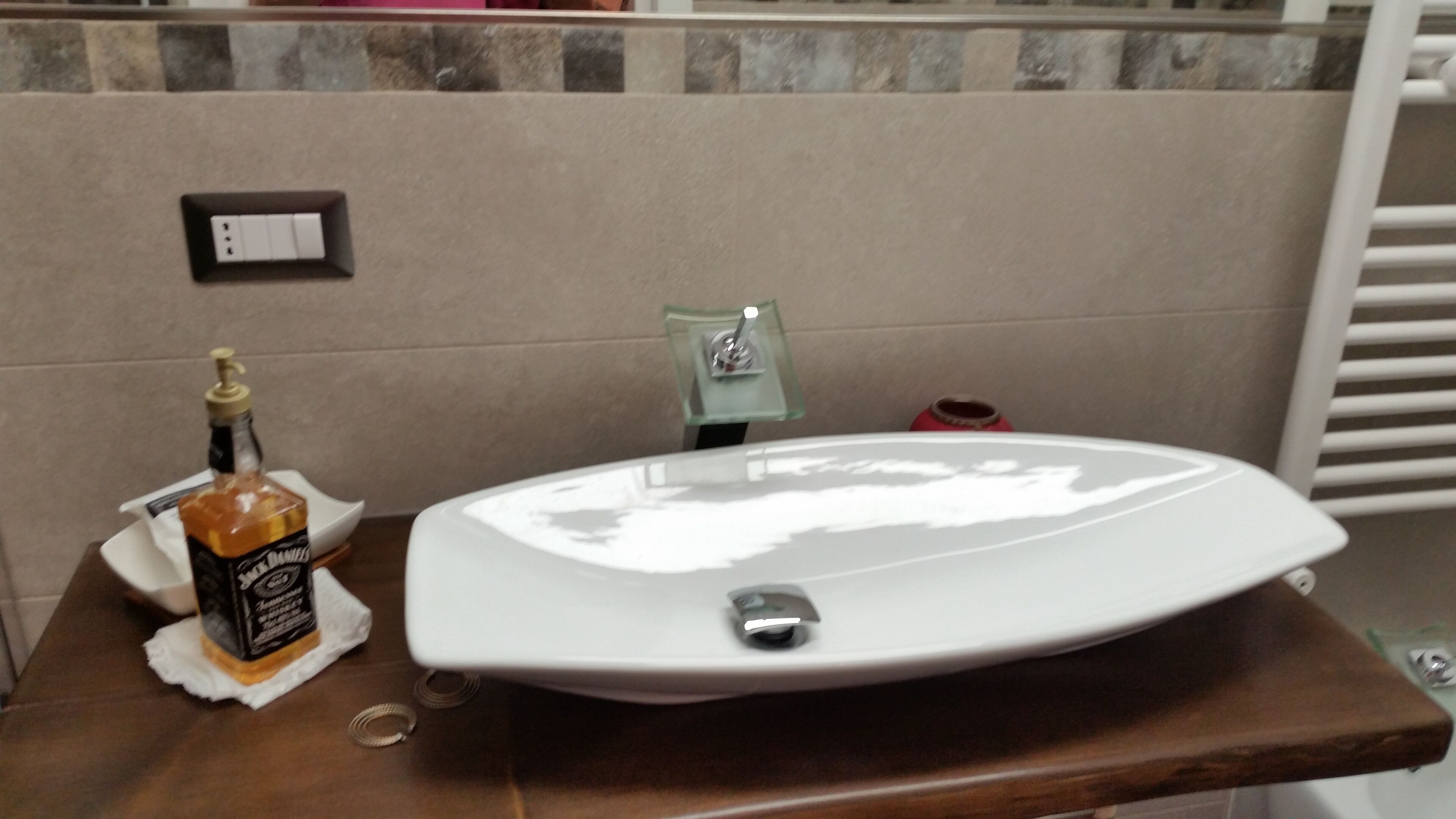 Un lavamanos de baño con un lavabo blanco, una botella de whisky Jack Daniel's y un dispensador de jabón de vidrio sobre una encimera de madera.