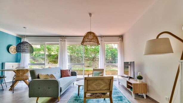 Ein modernes Wohnzimmer mit einer Couch, Sesseln, einem Fernseher und einem großen Fenster mit Blick ins Grüne.