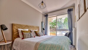 Ein ordentlich bezogenes Bett mit gemusterten Kissen, ein hölzerner Kopfteil und Blick ins Freie durch ein großes Fenster.