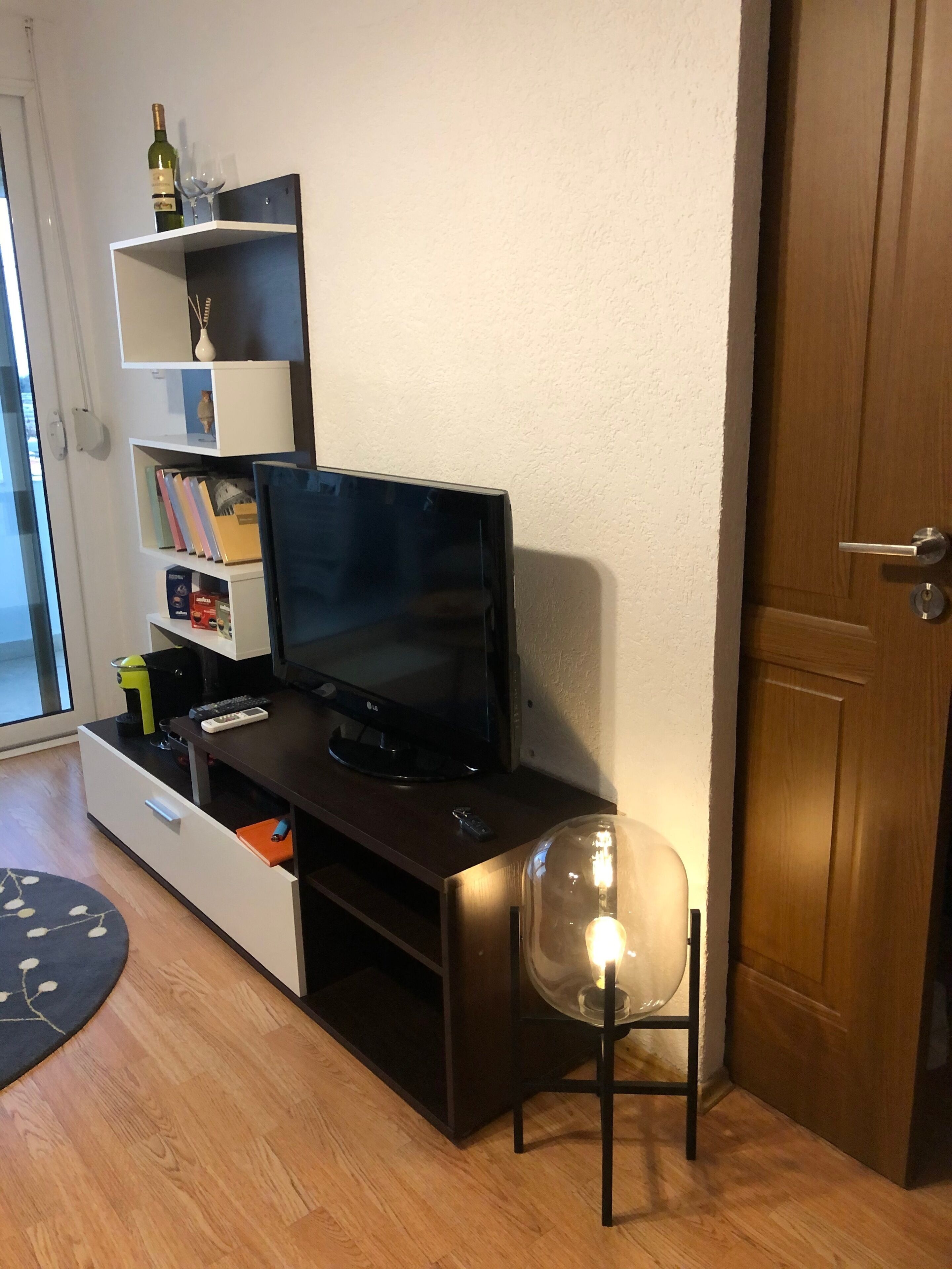 Ein kompakter Wohnbereich mit einem dunklen Holztisch, einem Flachbildfernseher, einem stuhlartigen Sitzmöbel mit Glastür und einem Bücherregal mit diversen Gegenständen.