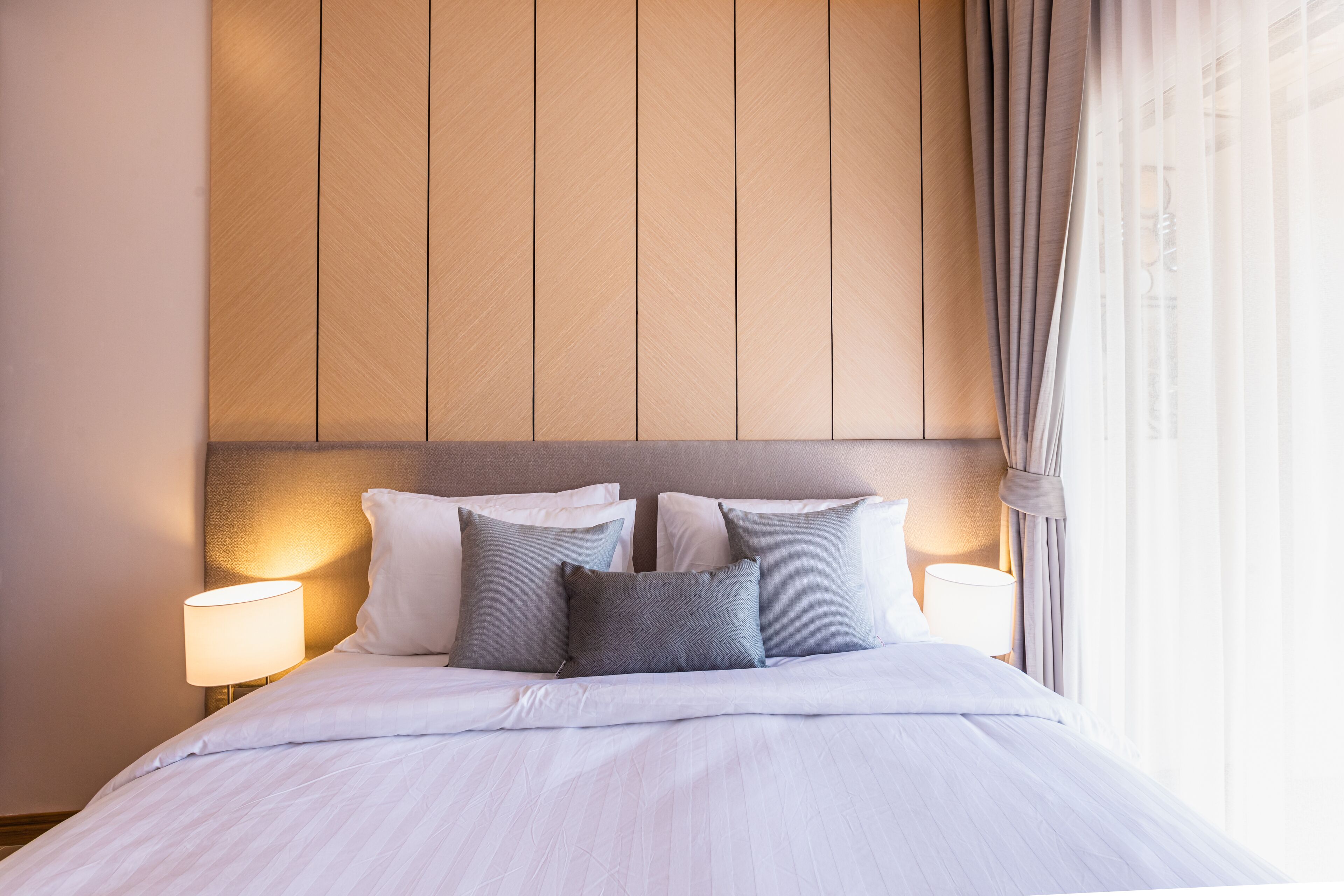 A neatly made bed with a headboard, two bedside lamps, and a window with curtains.