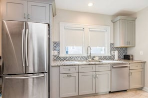 Una cocina con refrigerador de acero inoxidable, armarios blancos, salpicadero de azulejos y una ventana sobre el fregadero.