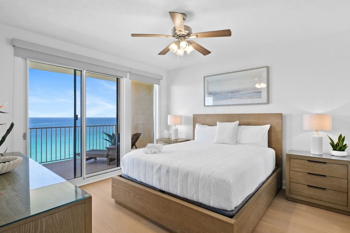 A bedroom with a bed, bedside tables, a ceiling fan, a painting, and a view of the ocean.