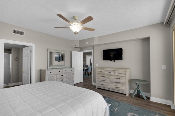 A bedroom with a bed, dresser, TV, ceiling fan, and a door leading to another room.