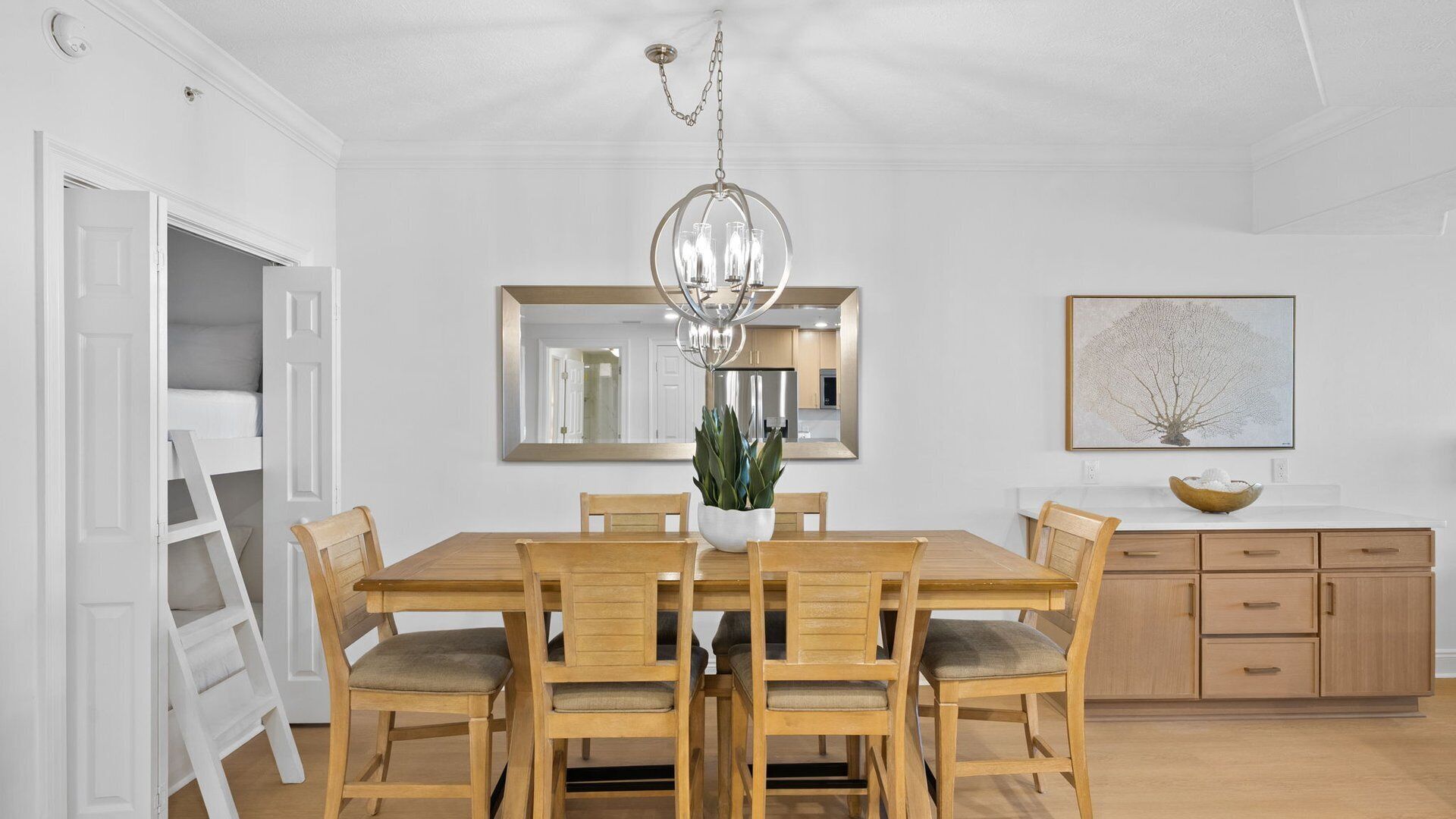A dining room with a wooden table and chairs, a chandelier, a potted plant, a large mirror, and a painting on the wall.