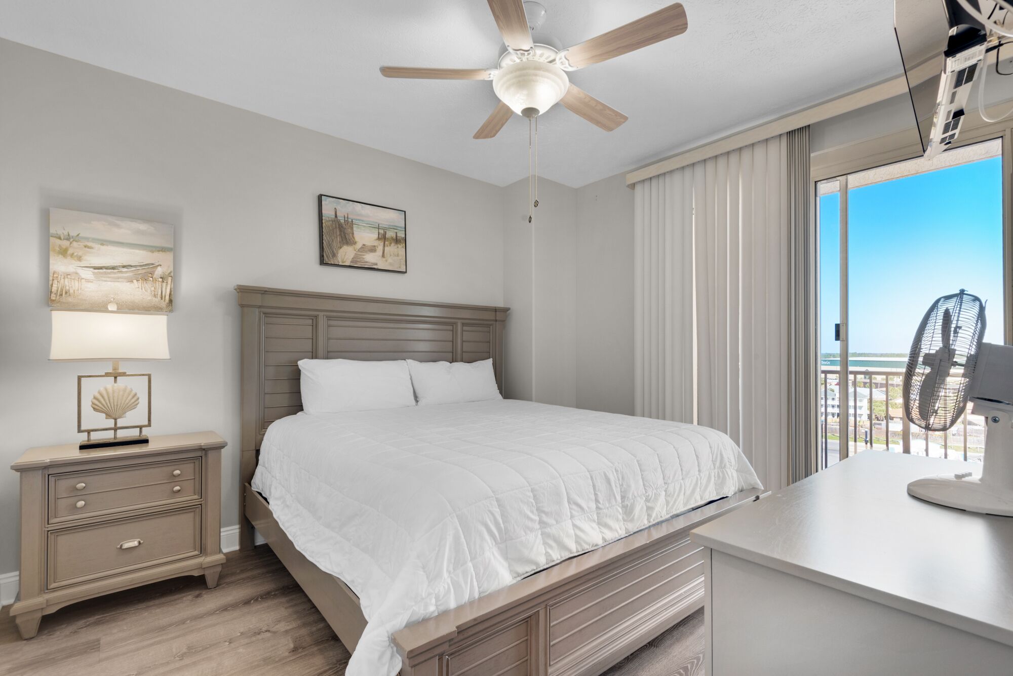 A bedroom with a bed, a nightstand, a ceiling fan, a painting, and a view of the outdoors.