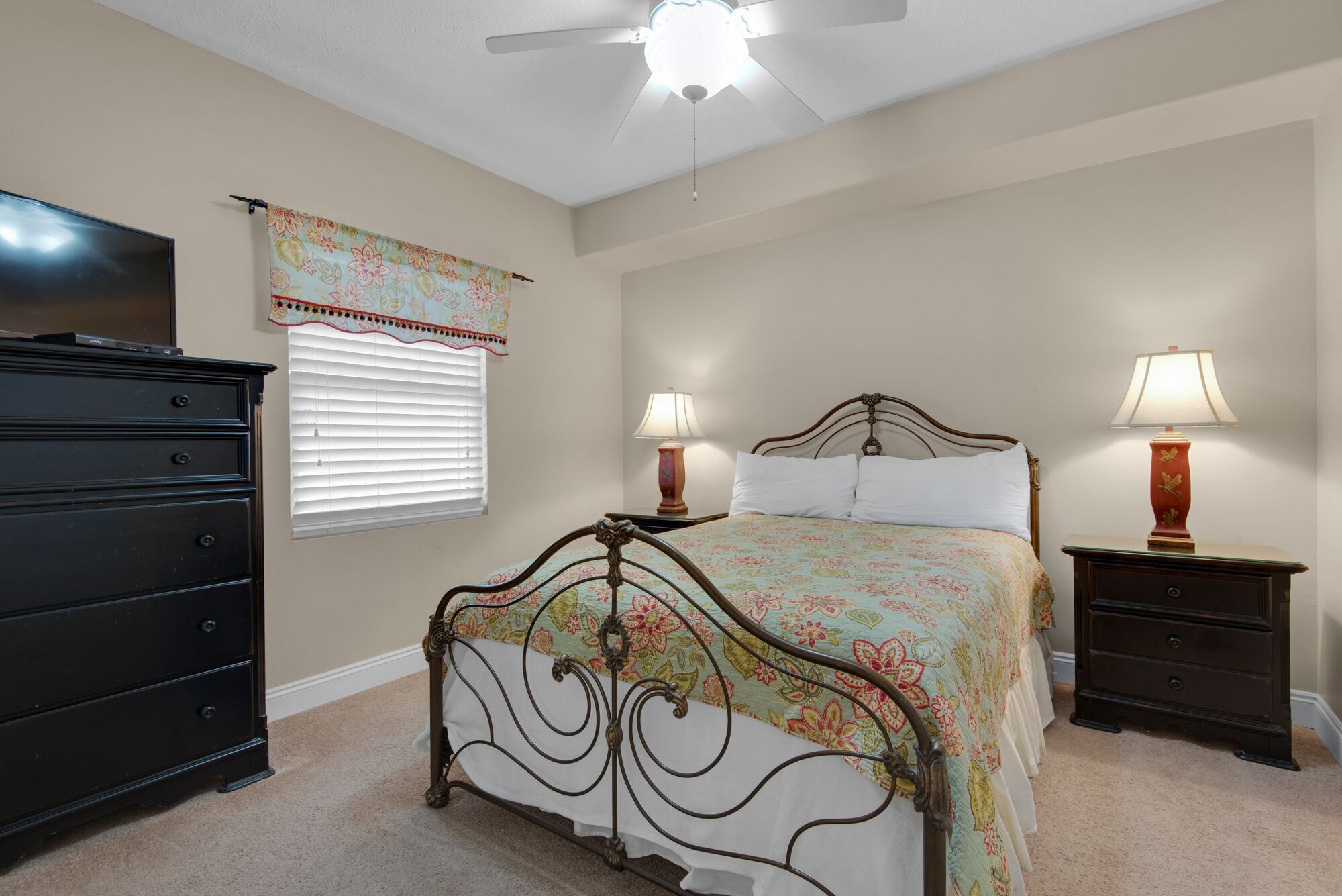 A bedroom with a bed, two bedside tables, a dresser, a ceiling fan, and a window with blinds.