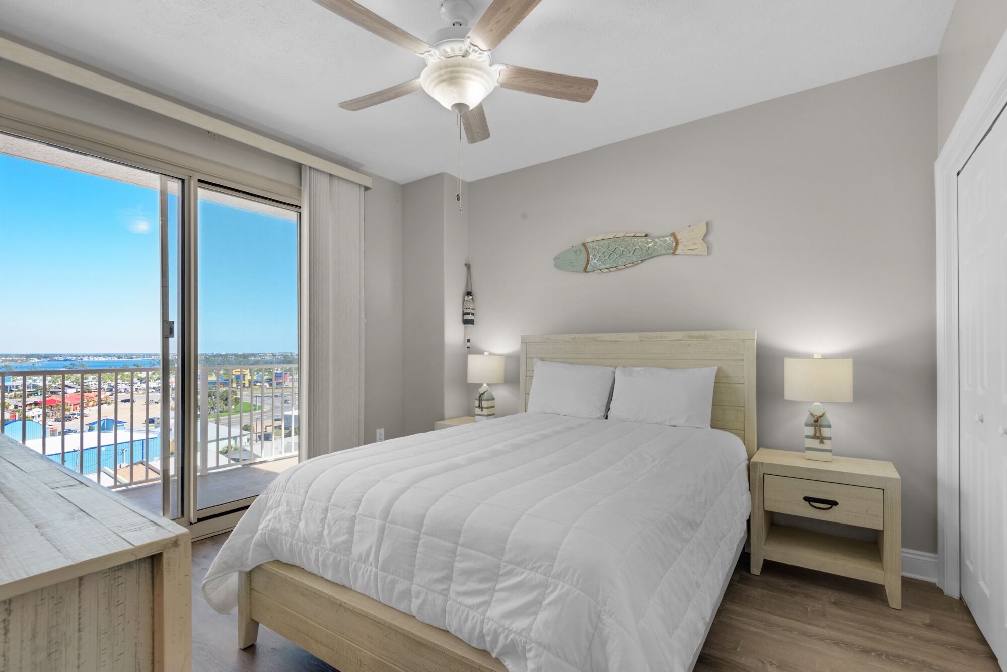 A bedroom with a large bed, a ceiling fan, a nightstand with a lamp, and a view of the city through a large window.