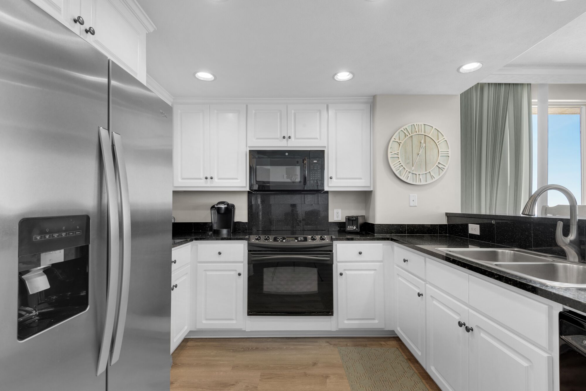 A modern kitchen with white cabinets, stainless steel appliances, a black countertop, and a large wall clock.