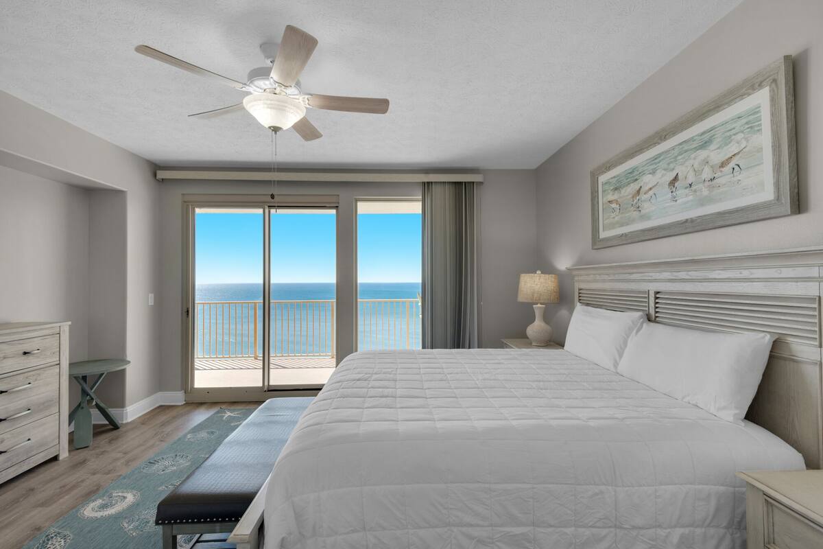 A hotel room with a large bed, a ceiling fan, a view of the ocean through the balcony doors, and a painting on the wall.