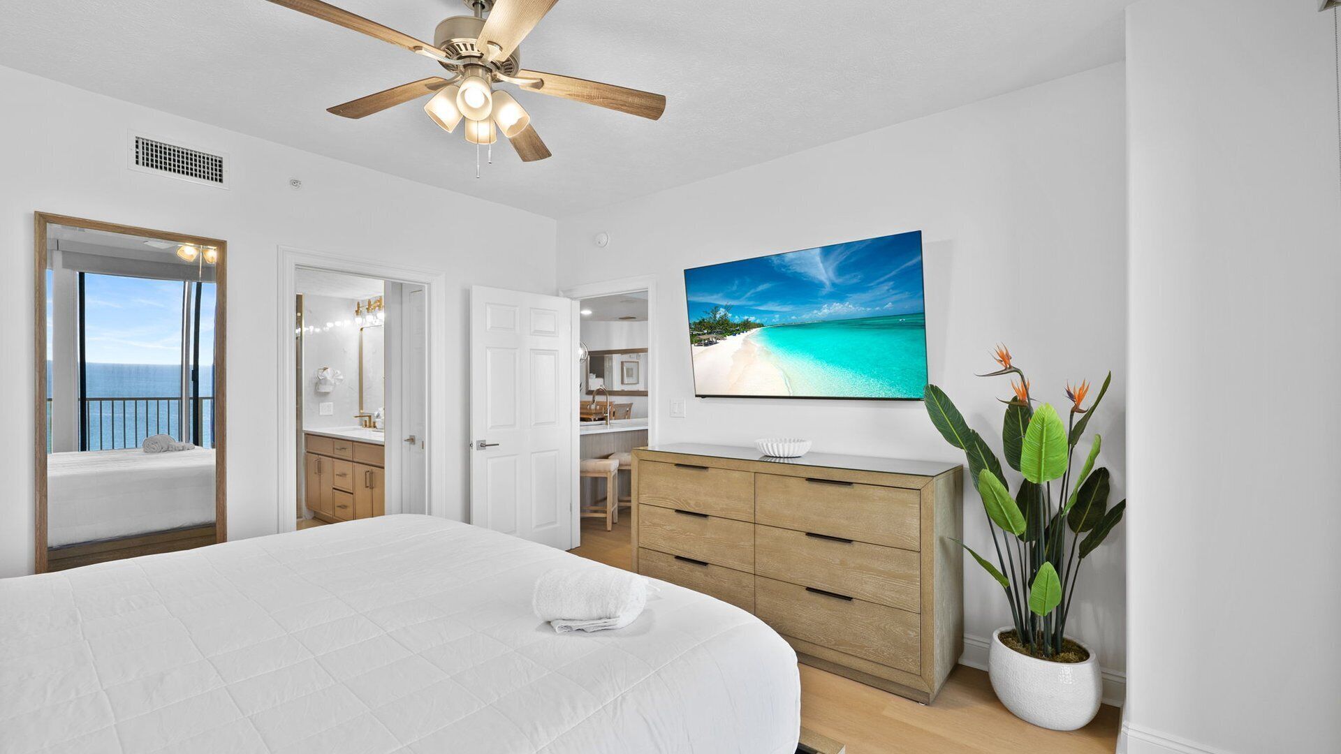 A bedroom with a large bed, a ceiling fan, a view of the sea through a window, a wooden dresser, and a potted plant.