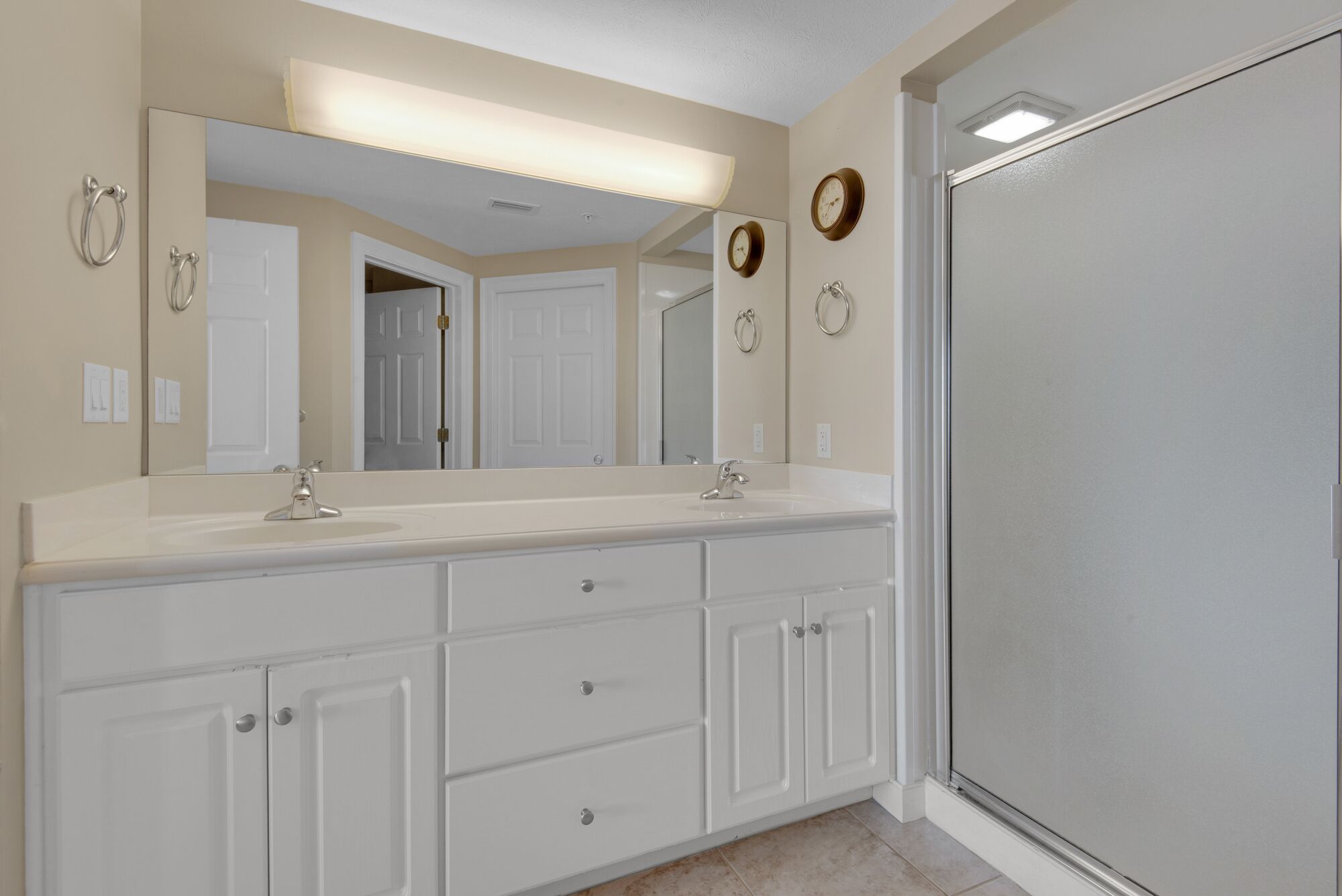 A bathroom with a double vanity, a large mirror, wall-mounted clocks, and a frosted glass shower door.