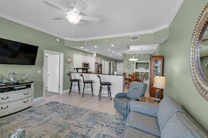 A spacious living room with a grey sofa, a coffee table, and a TV mounted on the wall. There is a kitchen area with white cabinets and a dining table with chairs.