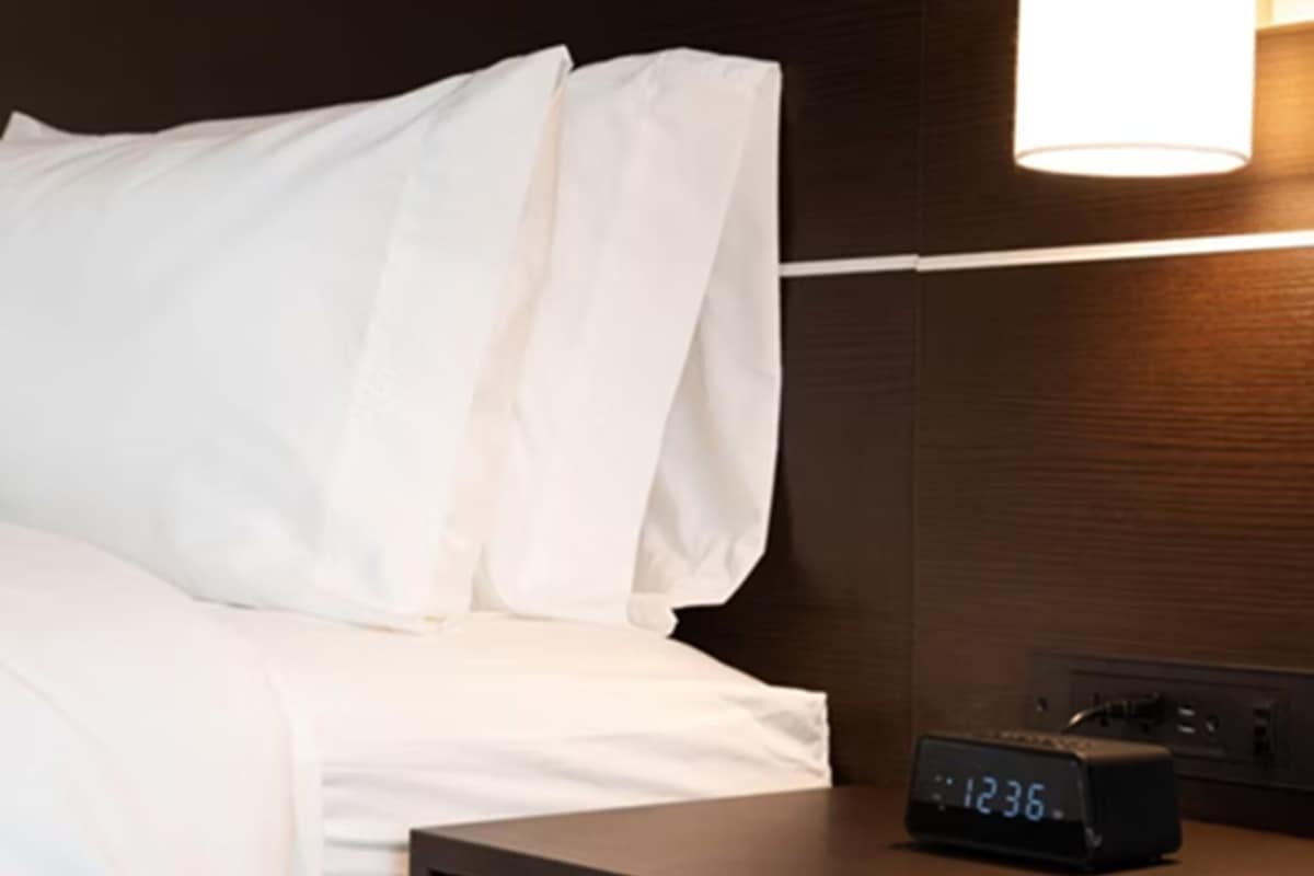 Habitació d'hotel amb llit, roba de llit blanca, una taula de nit amb rellotge digital i un llum fixada a la paret.