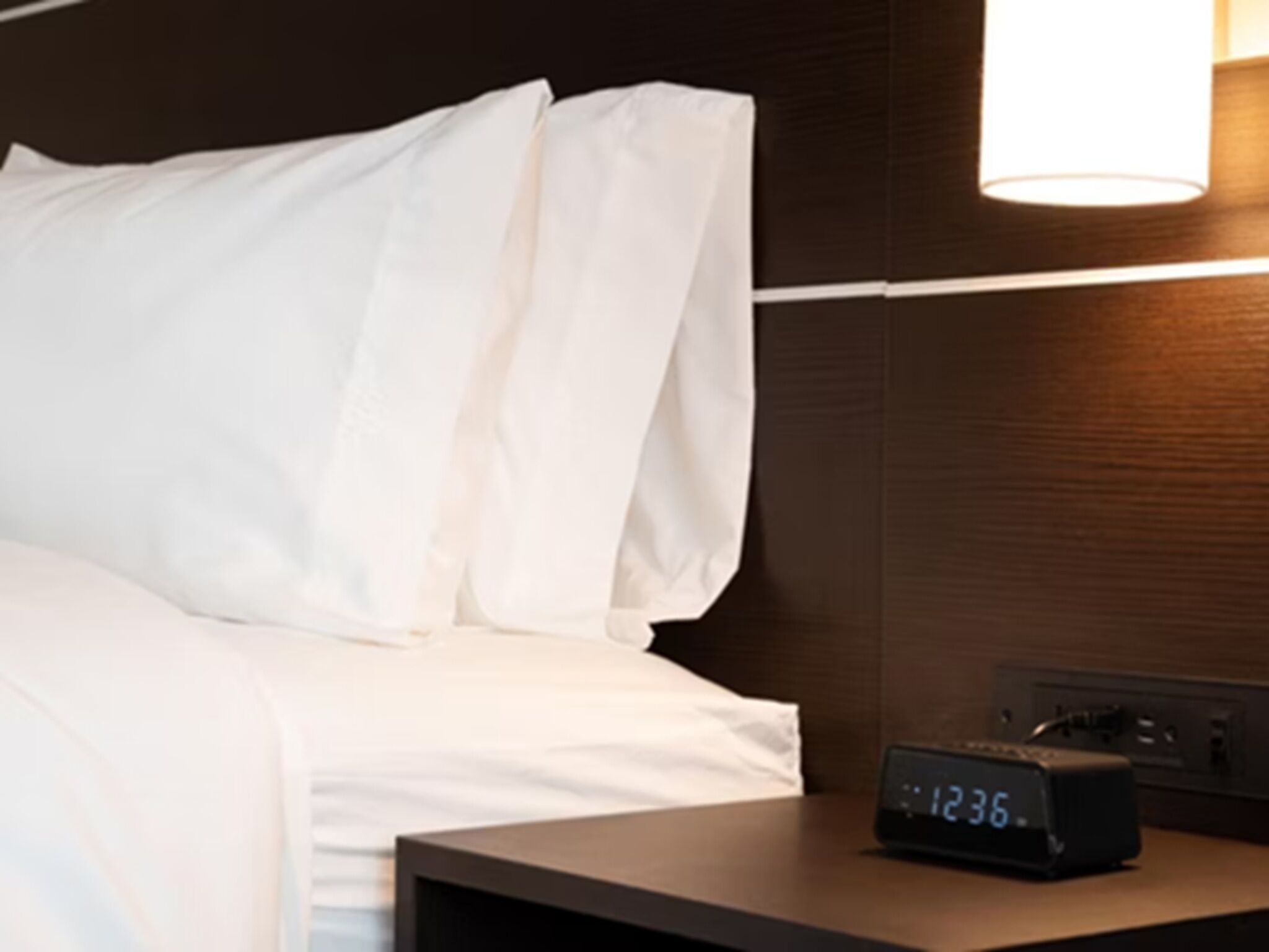 Habitación de hotel con cama, ropa de cama blanca, mesita de noche con reloj digital y lámpara de pared.