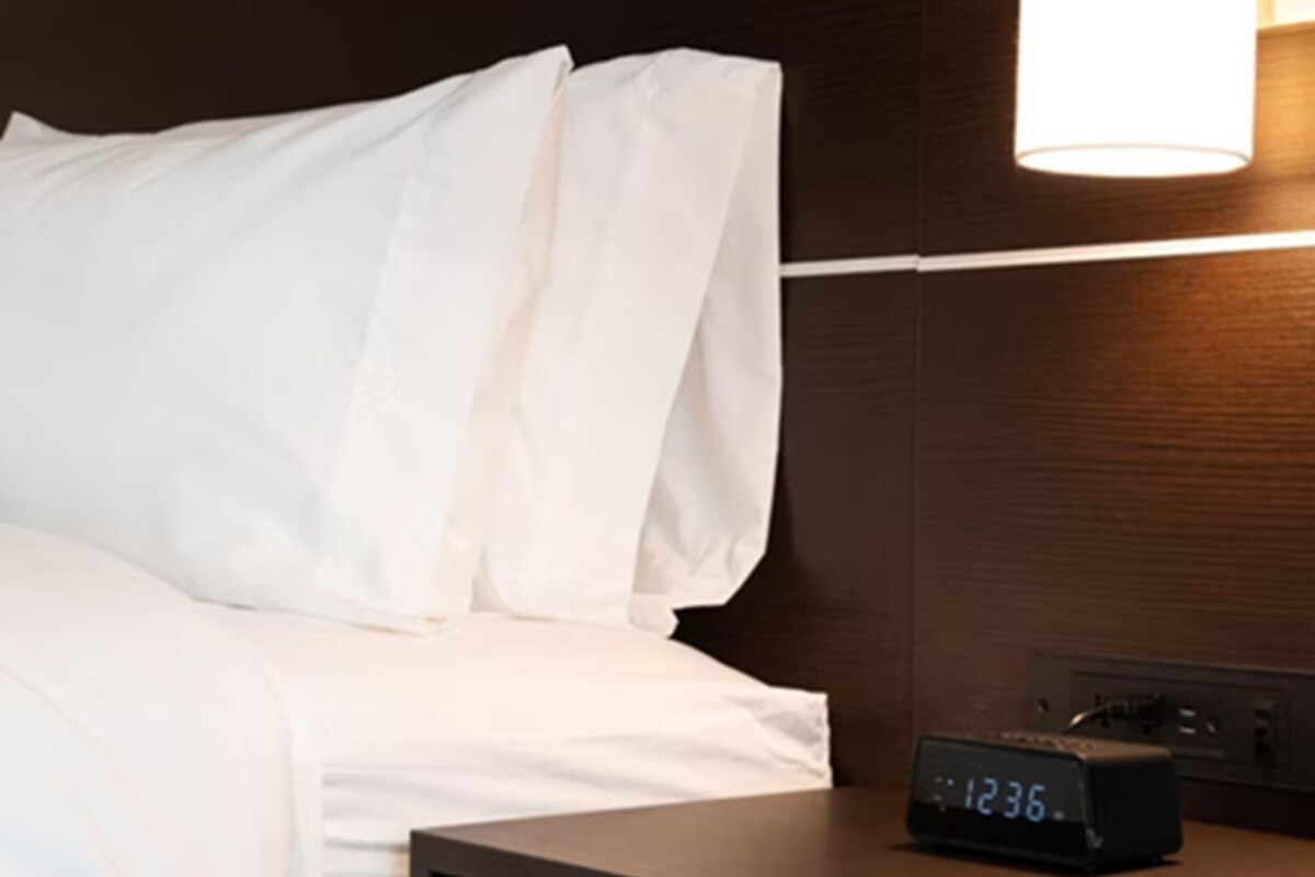 Habitació d'hotel amb llit, roba de llit blanca, una taula de nit amb rellotge digital i un llum fixada a la paret.