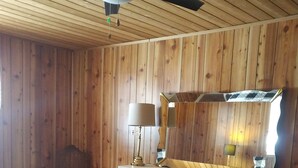 Ein Schlafzimmer mit Holzbalkendecke, einem Bett mit Steppdecke, einer Kommode mit Schubladen, einem Spiegel und einem Deckenventilator.