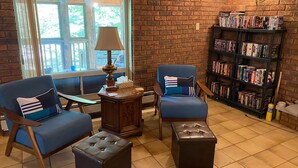 Eine gemütliche Leseecke mit zwei blauen Sesseln, einem kleinen Holztisch, einer Lampe, einem Bücherregal voller Bücher und Blick durch ein großes Fenster.