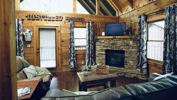 Interior de una cabaña de troncos con chimenea de piedra, un televisor montado encima y una zona de estar acogedora con sofá y mesa.