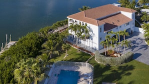 Un grand bâtiment blanc, surmonté d’un toit recouvert de tuiles rouges, entouré de palmiers et bordé d’une piscine.