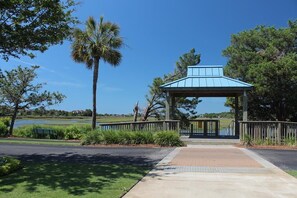 Un kiosque surmonté d’un toit bleu, entouré d’une clôture en bois, situé près d’une étendue d’eau et d’une végétation luxuriante.