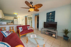 A living room with a red sofa, a glass coffee table, a TV on a stand, a ceiling fan, and a kitchen in the background.