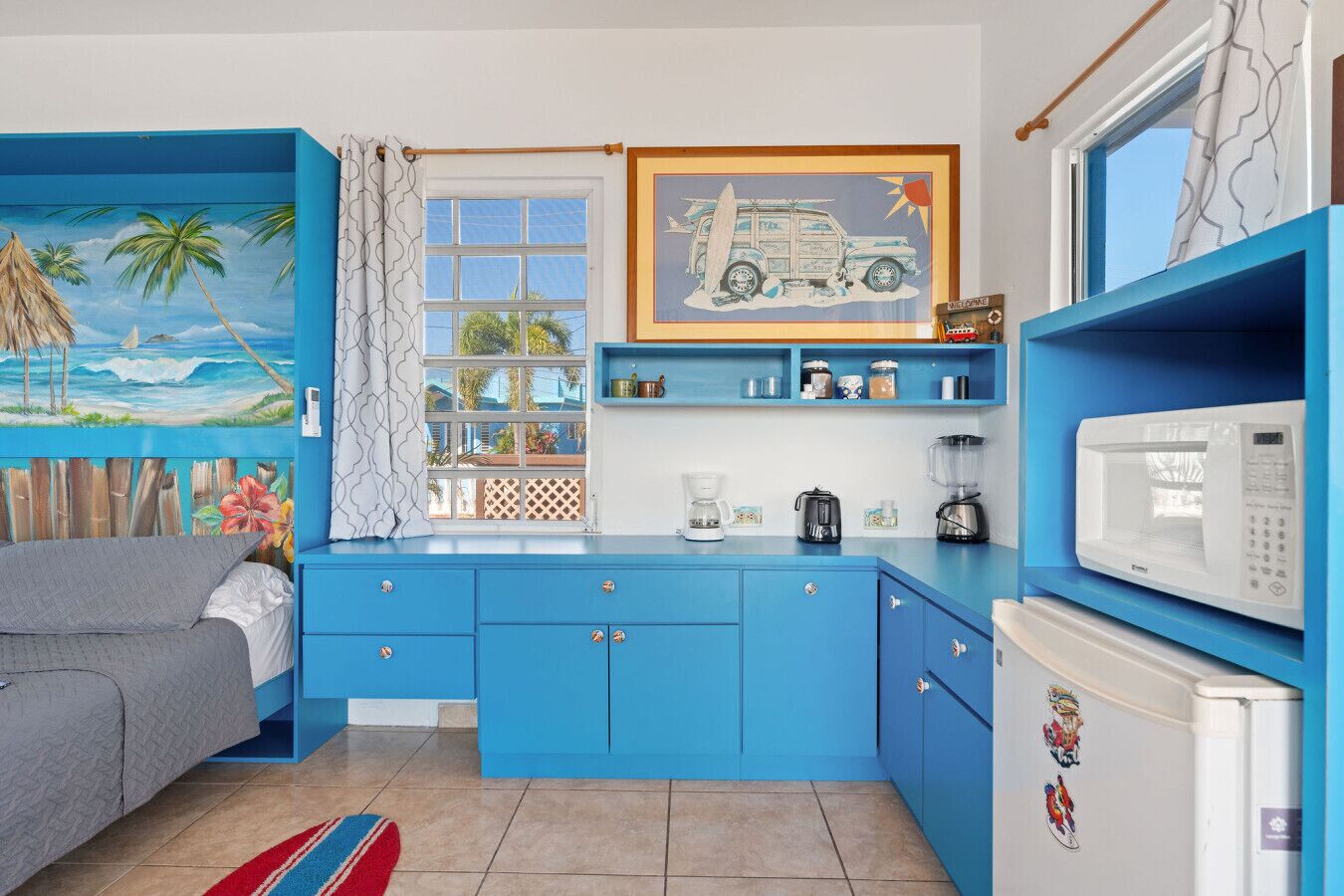 Een compacte kamer met een kitchenette, een bed en een raam met uitzicht op palmbomen en een strand.