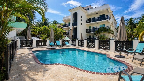 A rectangular swimming pool with a brick border, surrounded by lounge chairs and umbrellas, with a multi-story building in the background. - Ocean Terrace 202 a 4 min walk to Sandy Beach (Rincon)