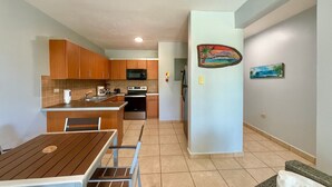 A compact kitchen with wooden cabinets, a microwave, and a stove. There is a dining table with chairs and a small island. The walls are painted light blue, and there is a painting on the wall. - Ocean Terrace 202 a 4 min walk to Sandy Beach (Rincon)