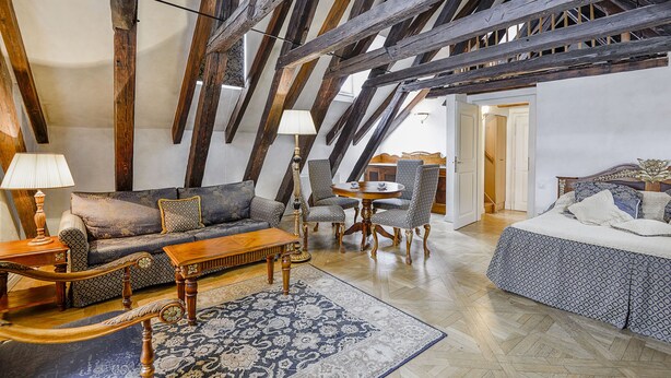 A loft-style room with exposed wooden beams, a sofa set, a round table, patterned chairs, a bed, and a patterned rug. - Maisonette 2 bedroom Apartment in the Heart of Prague (Prague)