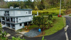 A two-story building with a flat roof, a covered parking area, and a small garden with a tree and a blue barrel. - Green Mountain Studio #1 (Rincón)