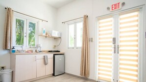 Cocina compacta con refrigerador, microondas y cafetera sobre la encimera, un lavamanos y un cubo de basura. Hay una puerta con un letrero de salida encima.