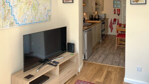 A modern kitchen with a flat-screen TV, a dining area with a red table and chairs, and a hallway leading to a living room. - 1 bedroom accommodation in Bowness (Bowness)