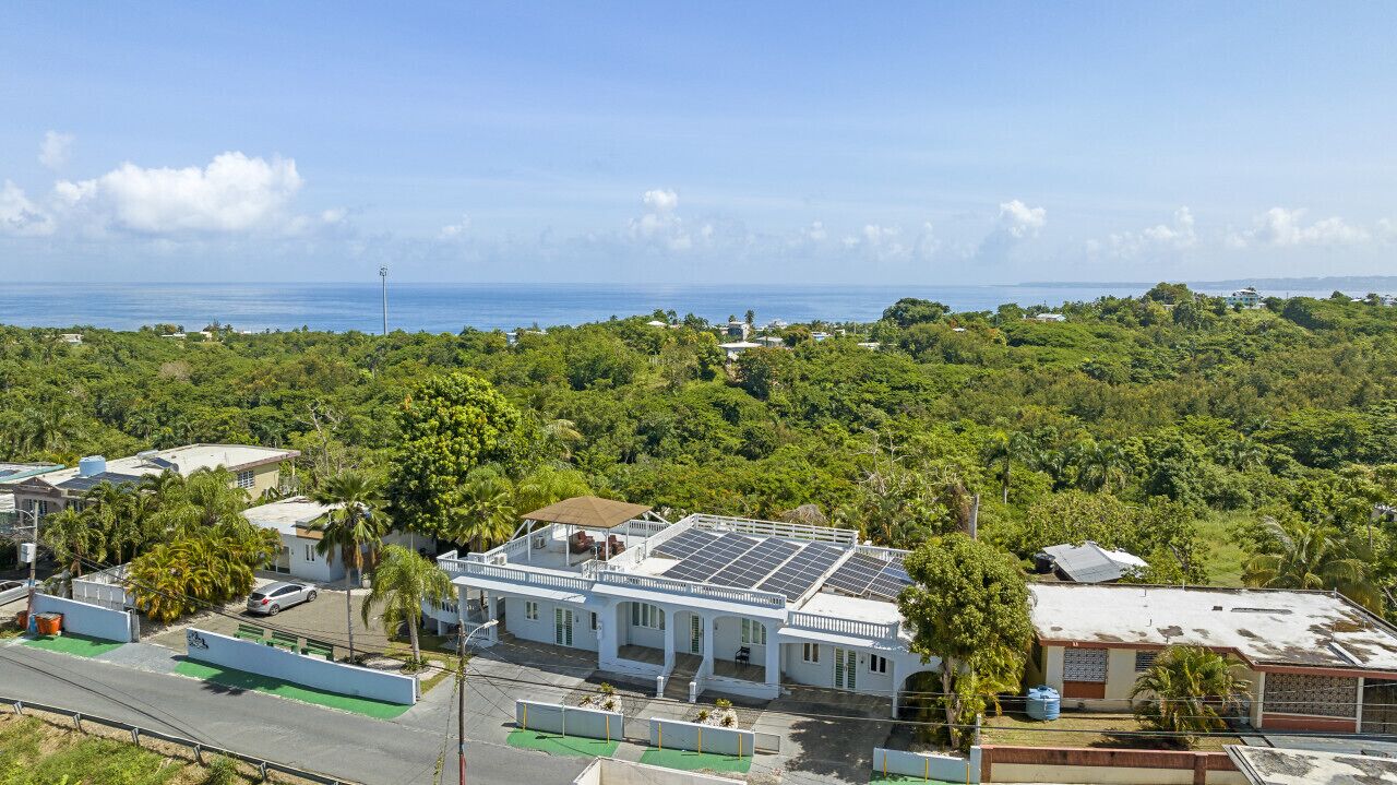 A coastal building with solar panels, surrounded by dense greenery and a clear view of the ocean.
