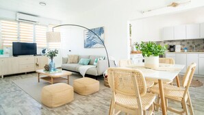 Een moderne woonkamer met een bank, salontafel, poefs en rieten stoelen. Er is een eettafel met stoelen, een televisie en een plant.
