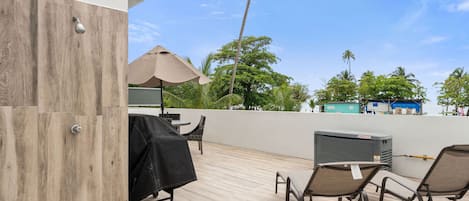 An outdoor patio with a grill, lounge chairs, and a view of palm trees and the sea.