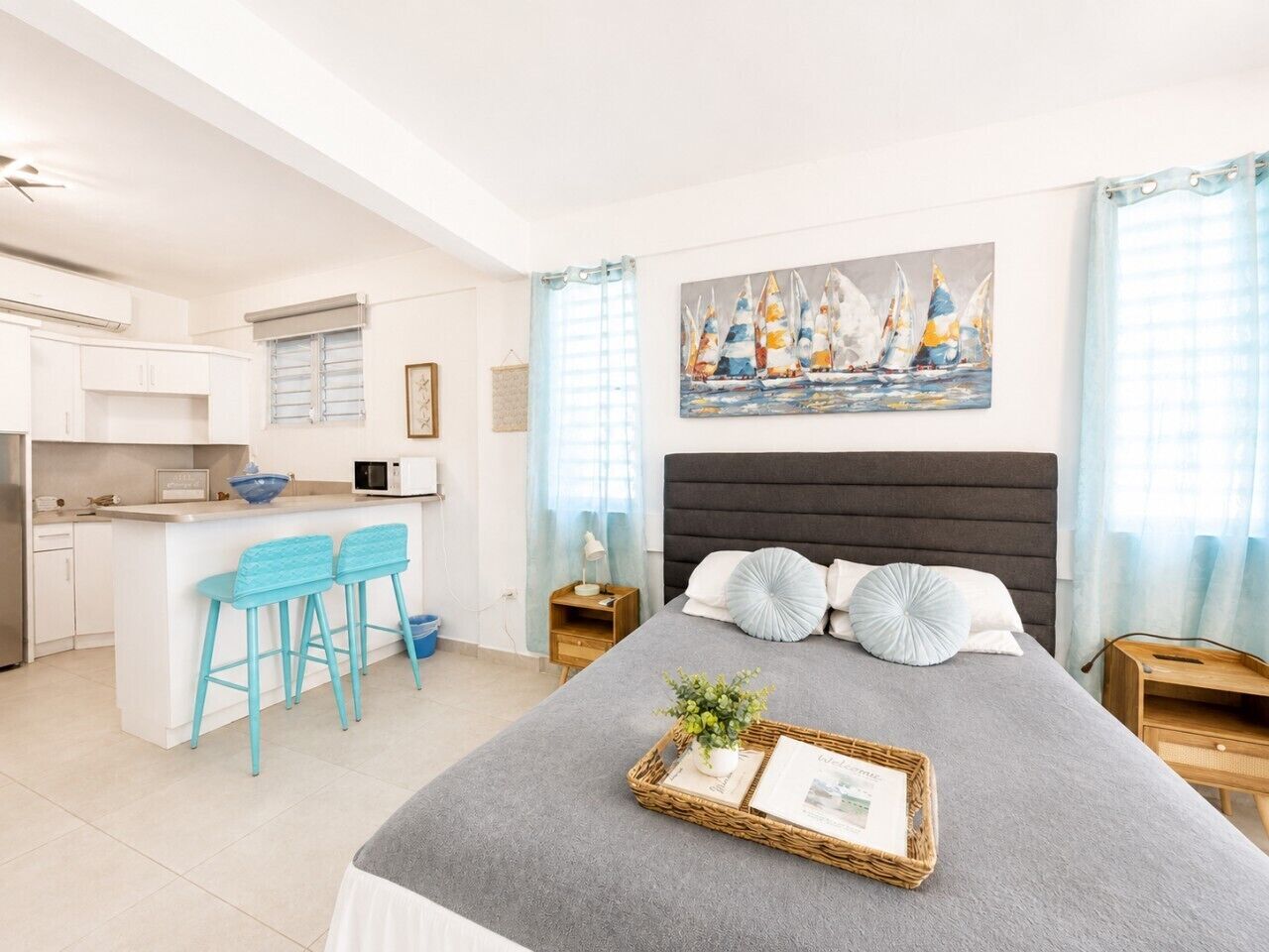 A modern hotel room with a bed, a small kitchenette, and a dining area with two blue bar stools.
