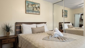 Een net opgemaakt bed met een beige sprei, kussens en een opgerolde handdoek. Een kleine plant staat op een nachtkastje naast het bed.