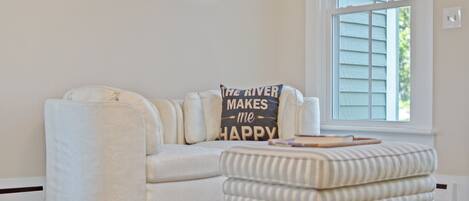 A living room with a white sofa, a striped ottoman, and a pillow with text. There are framed pictures on the wall and a window with blinds.