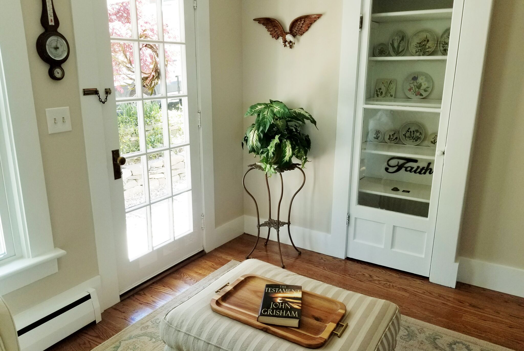 Une pièce avec un plancher en bois, une porte blanche, une fenêtre, une plante posée sur un support en métal, une bibliothèque ornée d’assiettes décoratives et un plateau sur lequel repose un livre intitulé « Le Testament » de John Grisham.